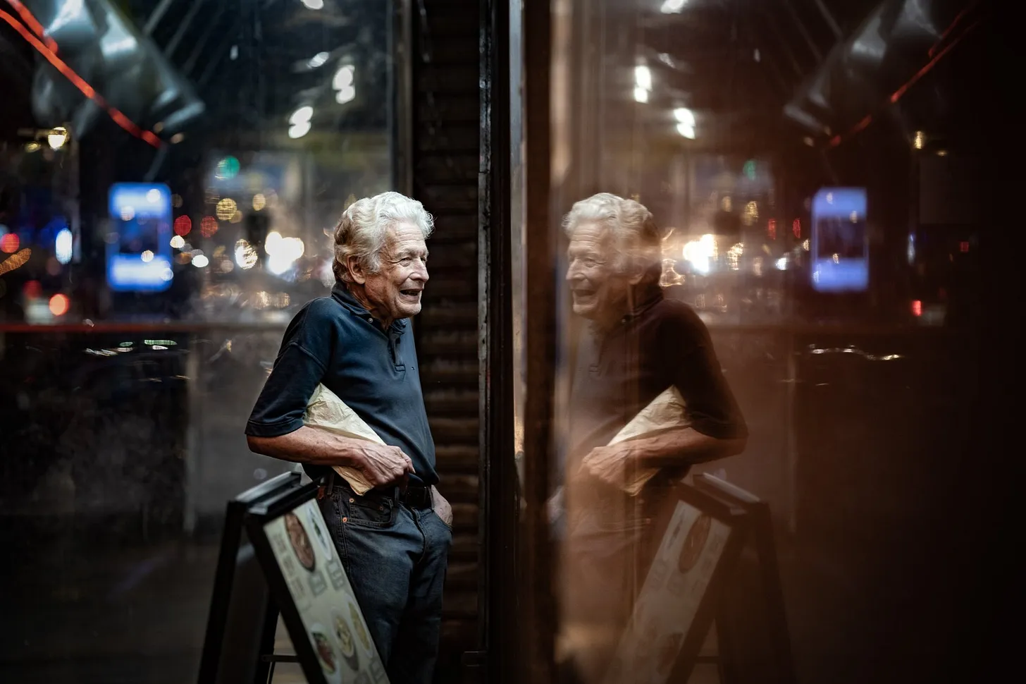 Photo of a man laughing into his reflection in a shop window at night.