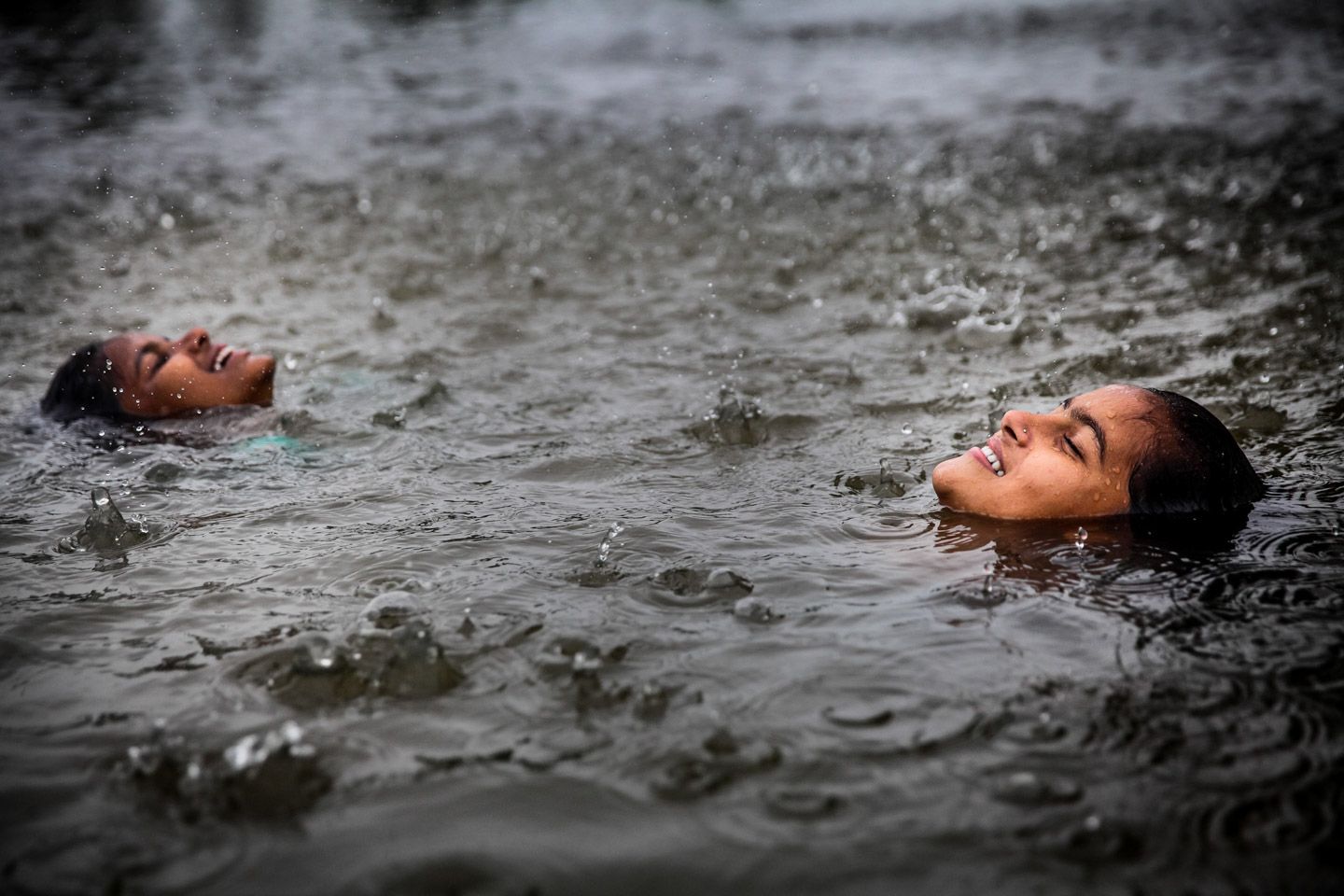 Two young girls in the water with only their faces showing, smiling, looking up into falling rain.