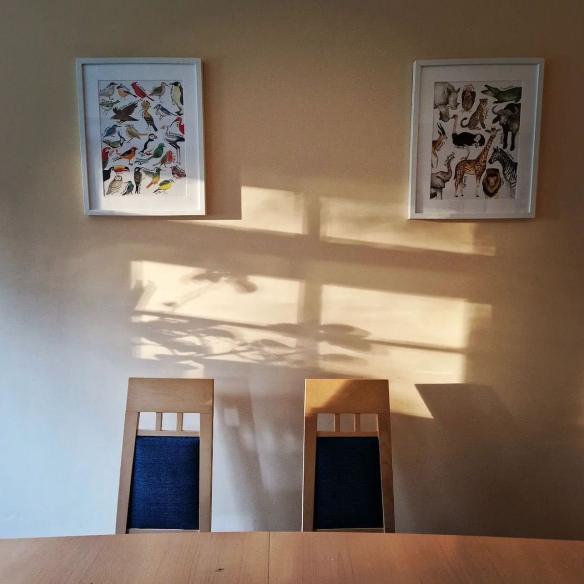 Plan shadows on a kitchen wall with two posters up with animals on them, and two dining chairs.