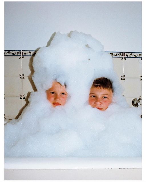 Vintage-looking photo of two young boys in a bath tub covered in bubbles, no smiles, having serious fun.